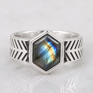 May include: A silver ring featuring a central hexagonal labradorite gemstone with blue and gold iridescence. The band is crafted from polished silver and displays a repeating chevron or arrow pattern engraved along the sides of the ring shank.