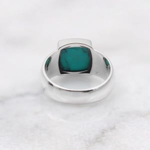 Peut inclure: Une bague en argent orn&eacute;e d'une pierre turquoise de taille carr&eacute;e avec des veines sombres. L'anneau est lisse et poli, pr&eacute;sentant une finition m&eacute;tallique qui contraste avec la couleur bleu-vert vive de la pierre sertie au centre.