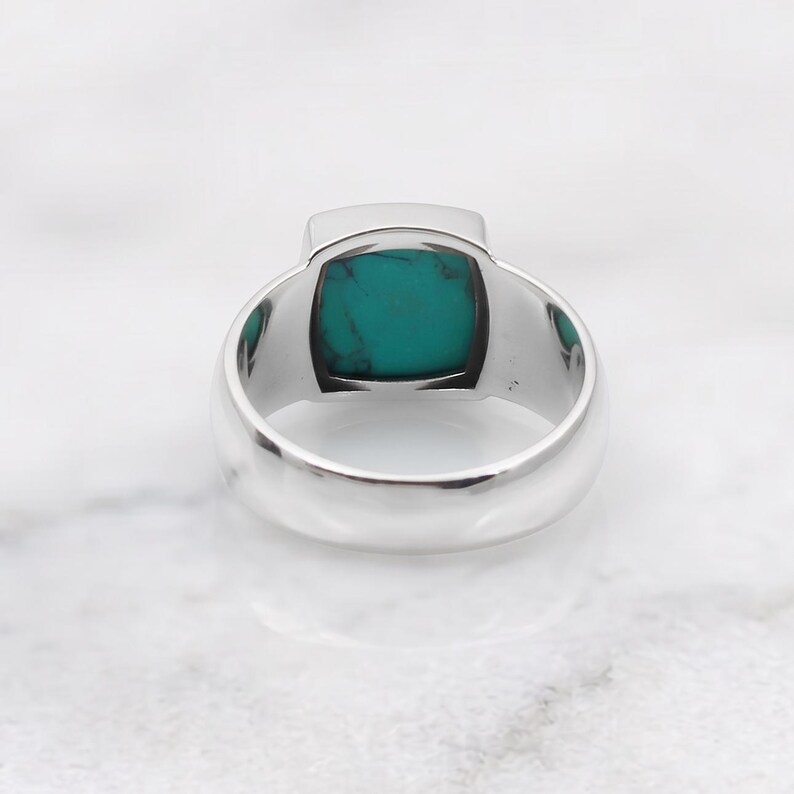 Peut inclure: Une bague en argent orn&eacute;e d'une pierre turquoise de taille carr&eacute;e avec des veines sombres. L'anneau est lisse et poli, pr&eacute;sentant une finition m&eacute;tallique qui contraste avec la couleur bleu-vert vive de la pierre sertie au centre.