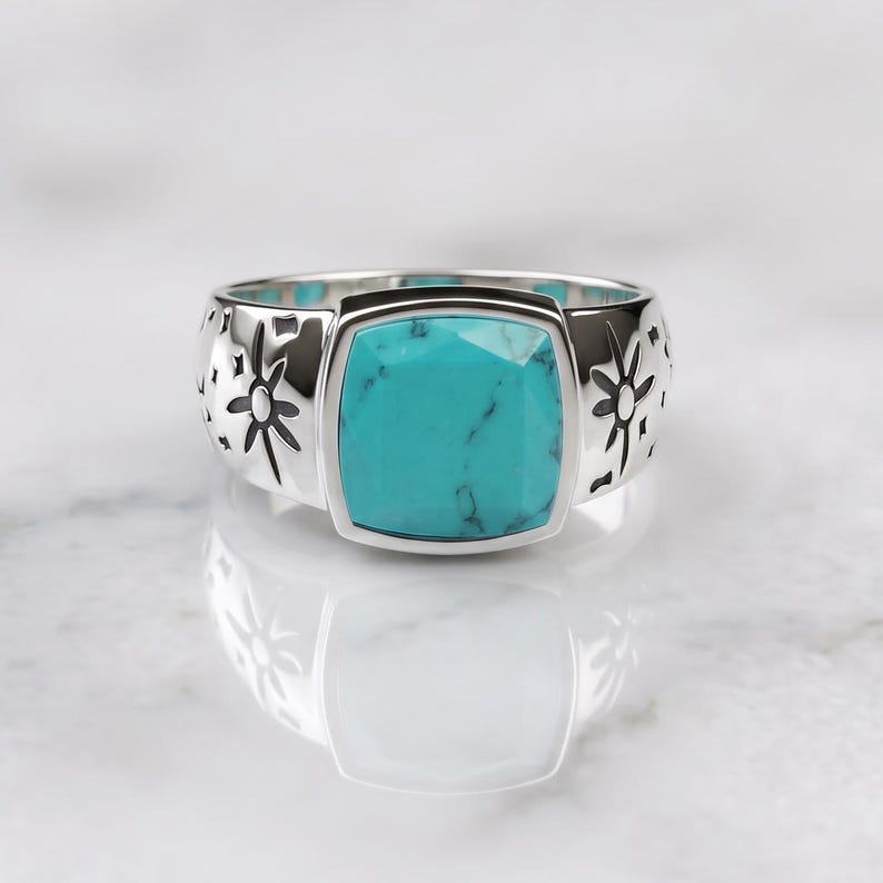 Peut inclure: Une bague argent&eacute;e orn&eacute;e d'une pierre turquoise carr&eacute;e &agrave; facettes avec des veines sombres. L'anneau est en m&eacute;tal poli et pr&eacute;sente des motifs grav&eacute;s en forme d'&eacute;toiles et de symboles c&eacute;lestes de chaque c&ocirc;t&eacute; de la pierre centrale.