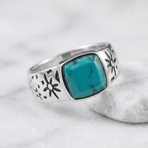 Peut inclure: Une bague en argent orn&eacute;e d'une pierre turquoise de taille carr&eacute;e avec des veines noires. L'anneau est grav&eacute; de motifs d'&eacute;toiles et de rayons de soleil de chaque c&ocirc;t&eacute; du sertissage central, cr&eacute;ant un design d&eacute;taill&eacute; sur la surface m&eacute;tallique.
