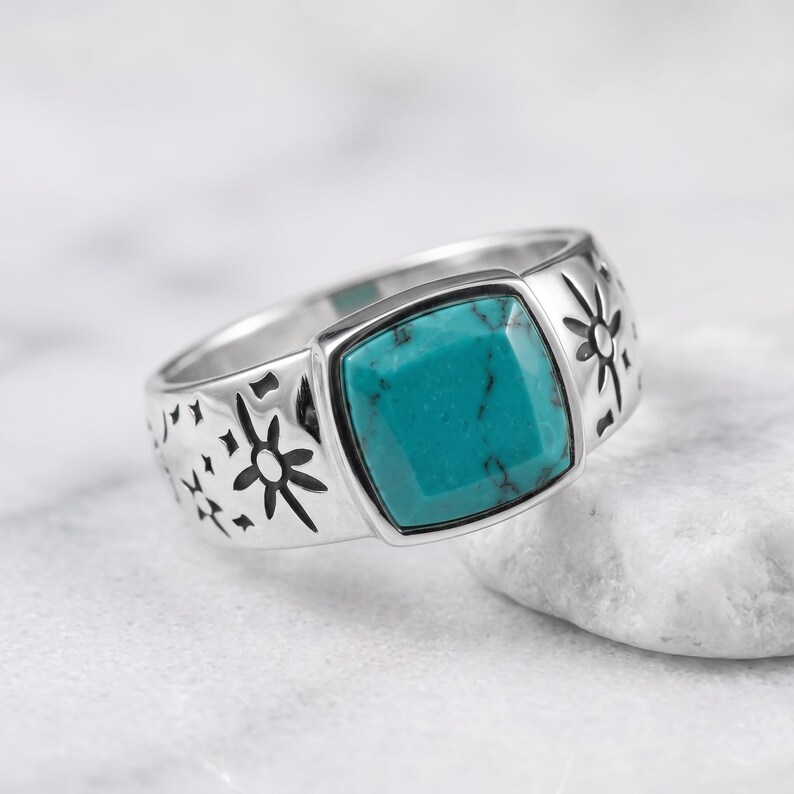 Peut inclure: Une bague en argent orn&eacute;e d'une pierre turquoise de taille carr&eacute;e avec des veines noires. L'anneau est grav&eacute; de motifs d'&eacute;toiles et de rayons de soleil de chaque c&ocirc;t&eacute; du sertissage central, cr&eacute;ant un design d&eacute;taill&eacute; sur la surface m&eacute;tallique.