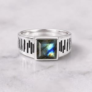 May include: A silver-toned ring featuring a square-cut labradorite gemstone with iridescent blue and yellow flashes. The band is crafted with a wide, flat profile and decorated with a series of vertical black rectangular cutouts along the sides.