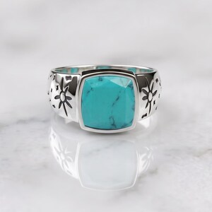 Peut inclure: Une bague argent&eacute;e orn&eacute;e d'une pierre turquoise carr&eacute;e &agrave; facettes avec des veines sombres. L'anneau est en m&eacute;tal poli et pr&eacute;sente des motifs grav&eacute;s en forme d'&eacute;toiles et de symboles c&eacute;lestes de chaque c&ocirc;t&eacute; de la pierre centrale.