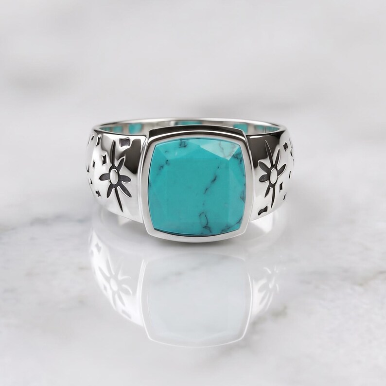 Peut inclure: Une bague argent&eacute;e orn&eacute;e d'une pierre turquoise carr&eacute;e &agrave; facettes avec des veines sombres. L'anneau est en m&eacute;tal poli et pr&eacute;sente des motifs grav&eacute;s en forme d'&eacute;toiles et de symboles c&eacute;lestes de chaque c&ocirc;t&eacute; de la pierre centrale.