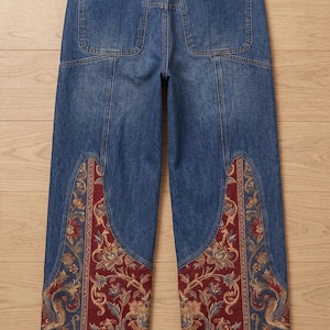 May include: Blue denim jeans featuring a high-waisted design with two back pockets, gold-toned rivets, and a blank black leather-like patch on the waistband. The lower legs are accented with wide, curved panels of red fabric with a floral pattern.