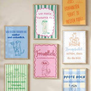 May include: A collection of seven rectangular wall art prints in light wooden frames, each featuring a meerkat illustration and German text. The designs include various patterns like stripes and checks in blue, green, pink, and yellow color palettes.