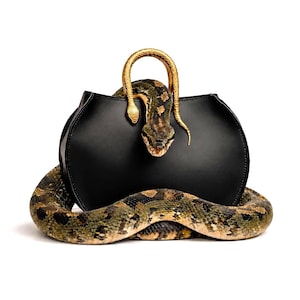 May include: A black handbag with a curved silhouette featuring a gold-coloured handle shaped like a snake. A large, patterned snake with green, brown, and black scales is coiled around the base of the bag and draped over the top handle.