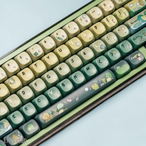 May include: A mechanical keyboard featuring a gradient of light to dark green keycaps. The keys are decorated with small illustrations of plants, leaves, flowers, bees, and frogs. The spacebar displays a detailed scene with a frog and foliage.