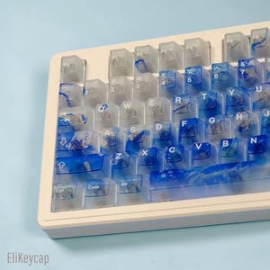 May include: A mechanical keyboard featuring translucent keycaps with a swirling blue and white ink-like pattern. The keys display standard QWERTY lettering and symbols in white. The keyboard frame is white, and the text EliKeycap is visible.