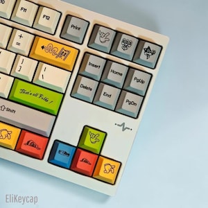 May include: A mechanical keyboard featuring a white frame and a variety of colorful keycaps. The keys include white, yellow, green, blue, and red colors with black graphic illustrations of cartoon hands, objects, and the text That's all Folks!