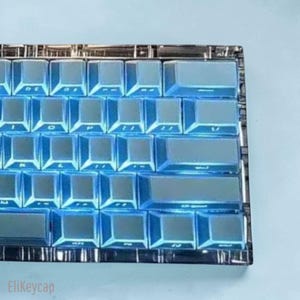 May include: A mechanical keyboard featuring translucent, light blue keycaps with a glossy finish. The keys are arranged in a standard layout with visible lettering on the surfaces. The frame of the keyboard is clear, revealing internal components.