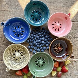 Può includere: Sei spremiagrumi in ceramica nei colori verde acqua, blu, rosa, marrone, crema e verde, ciascuno dotato di un cono centrale e di una piccola maniglia laterale. Un mucchio di mirtilli freschi si trova al centro, con fragole e fette di limone.