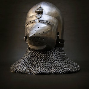 May include: A polished steel bascinet helmet featuring a pointed, perforated visor with horizontal eye slits and a decorative metal hinge at the top. The helmet is attached to a wide, flexible chainmail aventail that drapes around the base.