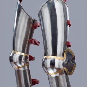 May include: Pair of polished steel leg armor with brass accents and red leather straps. The armor is designed with articulated knee protection and is likely for historical reenactment or display.