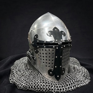 May include: A polished steel knight helmet featuring a pointed crown and a black metal cross overlay with fleur-de-lis accents. The faceplate includes a horizontal eye slit and a perforated lower section, resting on a circular base of chainmail armour.