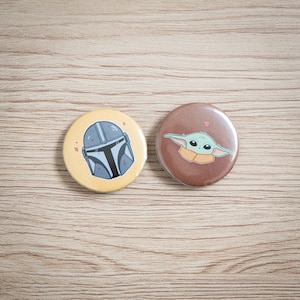 Peut inclure: Deux badges ronds représentant des personnages de Star Wars. L'un a un fond jaune avec un design de casque Mandalorien. L'autre a un fond marron avec un design de personnage Grogu.