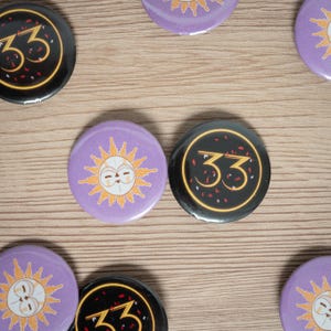 Peut inclure: Plusieurs badges ronds. Certains sont noirs avec le chiffre "33" en or, entouré de confettis. D'autres sont lavande avec un motif de soleil. Le soleil a un visage et des rayons.