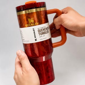 May include: A 40 oz Stanley The Flowstate Quencher Tumbler in a deep red and orange marbled pattern. It features a metallic gold rim, an orange handle, and a dark red lid with an orange tab. A white product label displays the Stanley logo and text.