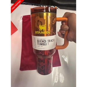 May include: A metallic orange and red Stanley Flowstate Quencher H2.0 tumbler with a floral pattern, a large orange handle, and a clear lid. The label reads The Flowstate Quencher H2.0 Tumbler 40 oz | 1.18 L. A dark red fabric pouch is visible behind.