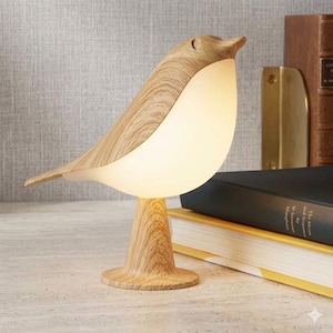 May include: A bird-shaped table lamp featuring a light wood grain finish on the head, tail, and base, with a smooth, opaque, off-white glowing body that emits a soft light. The lamp stands upright on a circular base, showcasing a clean design.