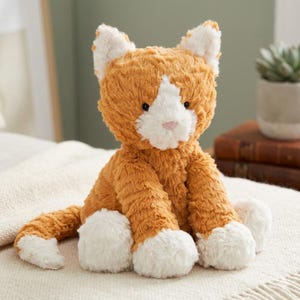Peut inclure: Une peluche chat orange et blanc, avec un nez rose et des yeux noirs. Le chat a des pattes et des oreilles blanches. La peluche est posée sur une couverture crème.