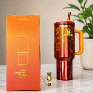 May include: A 40 oz Stanley Quencher H2.0 tumbler in a metallic orange finish with a floral pattern, a matching orange handle, and a straw. Beside it is an orange box with the text The FlowState Quencher H2.0 Tumbler 40 oz | 1.18 L and a small gold pineapple charm.