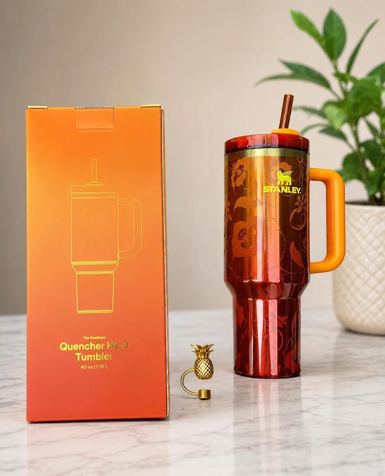 May include: A 40 oz Stanley Quencher H2.0 tumbler in a metallic orange finish with a floral pattern, a matching orange handle, and a straw. Beside it is an orange box with the text The FlowState Quencher H2.0 Tumbler 40 oz | 1.18 L and a small gold pineapple charm.