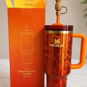 May include: A 40 oz Stanley Karol G Quencher H2.0 tumbler in metallic orange with a floral pattern and gold logo. It features an orange handle, a dark red lid, and a straw topped with a gold pineapple charm, standing next to its orange box.