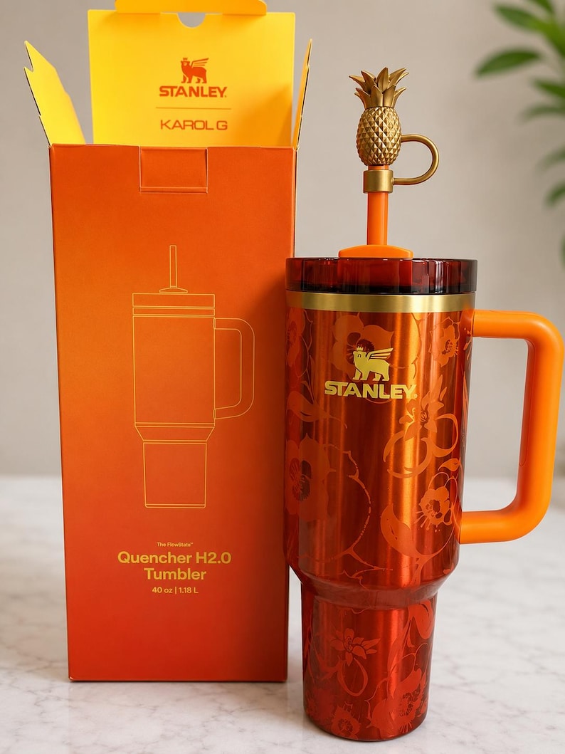 May include: A 40 oz Stanley Karol G Quencher H2.0 tumbler in metallic orange with a floral pattern and gold logo. It features an orange handle, a dark red lid, and a straw topped with a gold pineapple charm, standing next to its orange box.