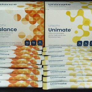 Op de afbeelding: Twee rechthoekige dozen met Unicity Balance en Unimate voedingssupplementen met meerdere individuele stickverpakkingen ervoor. De Balance-doos is wit met oranje cirkels, terwijl de Unimate-doos wit is met gele organische vormen.
