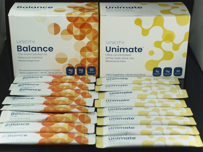 Op de afbeelding: Twee rechthoekige dozen met Unicity Balance en Unimate voedingssupplementen met meerdere individuele stickverpakkingen ervoor. De Balance-doos is wit met oranje cirkels, terwijl de Unimate-doos wit is met gele organische vormen.