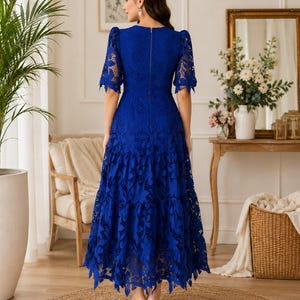 May include: A royal blue midi dress featuring an intricate floral lace overlay, short puff sleeves, and a tiered skirt. The dress has a fitted bodice with a visible vertical zipper closure along the center back and a scalloped hemline.