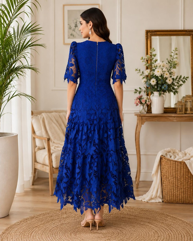 May include: A royal blue midi dress featuring an intricate floral lace overlay, short puff sleeves, and a tiered skirt. The dress has a fitted bodice with a visible vertical zipper closure along the center back and a scalloped hemline.