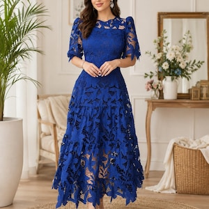 May include: A royal blue midi dress featuring intricate floral lace detailing throughout. The garment has short puff sleeves, a round neckline, and a tiered skirt design with a scalloped hemline. The lace pattern is dense and covers the entire dress.