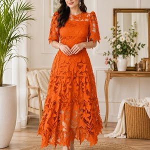 May include: A vibrant orange midi dress featuring intricate floral lace detailing throughout. The garment has short puff sleeves with tie accents, a round neckline, and a tiered skirt that falls to a mid-calf length with a scalloped hemline.