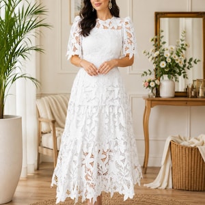 May include: A white midi-length dress featuring intricate floral lace detailing, short puff sleeves, a round neckline, and a tiered skirt with a scalloped hemline. The garment is constructed from a semi-sheer lace fabric with a solid white lining.