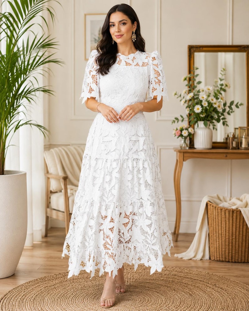 May include: A white midi-length dress featuring intricate floral lace detailing, short puff sleeves, a round neckline, and a tiered skirt with a scalloped hemline. The garment is constructed from a semi-sheer lace fabric with a solid white lining.