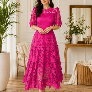 May include: A vibrant fuchsia midi dress featuring intricate floral lace detailing, short puff sleeves, a round neckline, and a tiered skirt with a scalloped hemline. The garment is constructed with a solid-colored lining beneath the lace overlay.