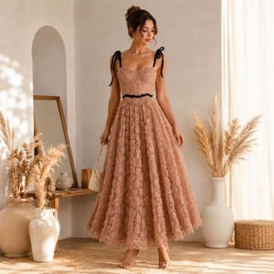 May include: A dusty rose midi dress featuring a textured floral lace overlay, a sweetheart neckline, and thin black shoulder straps. The bodice is fitted with a black ribbon waistband, and the skirt flows into a full, A-line silhouette.