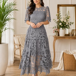 May include: A slate gray midi dress featuring intricate floral lace overlay, short puff sleeves, a round neckline, and a tiered skirt. The garment is constructed with a solid lining underneath the lace, creating a layered texture throughout the bodice and skirt.
