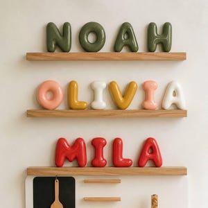 Peut inclure: Trois étagères en bois présentent des lettres en céramique style ballon. L'étagère supérieure affiche NOAH en vert olive. L'étagère du milieu affiche OLIVIA en pêche, jaune, blanc, jaune, pêche et blanc. L'étagère inférieure affiche MILA en rouge.