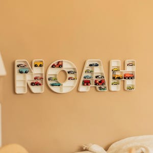 K&ouml;nnte beinhalten: Eine an der Wand befestigte, cremefarbene Holznamensanzeige mit der Aufschrift "NOAH". Jeder Buchstabe ist ein Regal, das mit Miniatur-Spielzeugautos in verschiedenen Farben gef&uuml;llt ist. Die Anzeige ist an einer hellbraunen Wand angebracht.