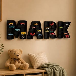 K&ouml;nnte beinhalten: Eine schwarze, an der Wand befestigte Anzeige mit der Aufschrift "MARK" mit einzelnen F&auml;chern f&uuml;r Miniatur-Spielzeugautos in verschiedenen Farben. Ein Teddyb&auml;r, ein Kissen und eine gr&uuml;ne Decke befinden sich auf einem Holzregal darunter. Ein gerahmtes Bild eines gelben Jeeps h&auml;ngt an der Wand.