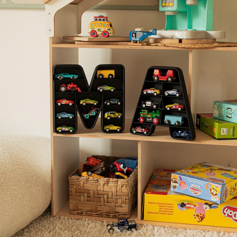 K&ouml;nnte beinhalten: Ein Holzregal mit Spielzeugautos, die in schwarzen, buchstabenf&ouml;rmigen Organisatoren ausgestellt sind. Die Autos sind in verschiedenen Farben und Stilen gehalten. Weitere Spielzeuge, darunter ein Bus, ein Hubschrauber und Knetboxen, sind ebenfalls zu sehen.