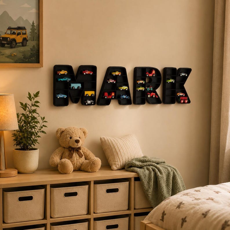 K&ouml;nnte beinhalten: Eine schwarze, an der Wand befestigte Anzeige mit der Aufschrift "MARK" mit einzelnen F&auml;chern f&uuml;r Miniatur-Spielzeugautos in verschiedenen Farben. Ein Teddyb&auml;r, ein Kissen und eine gr&uuml;ne Decke befinden sich auf einem Holzregal darunter. Ein gerahmtes Bild eines gelben Jeeps h&auml;ngt an der Wand.