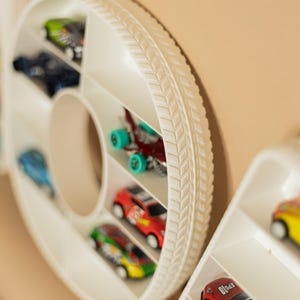 K&ouml;nnte beinhalten: Ein wei&szlig;er, reifenf&ouml;rmiger Spielzeugauto-Organizer mit mehreren F&auml;chern, in denen jeweils ein farbenfrohes Spielzeugauto untergebracht ist. Der Organizer hat ein strukturiertes Reifenprofil-Design am &auml;u&szlig;eren Rand. Die Spielzeugautos sind in verschiedenen Farben.