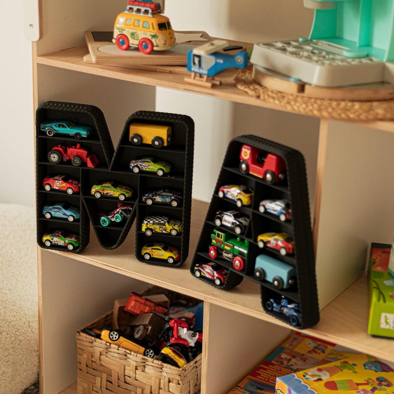 K&ouml;nnte beinhalten: Schwarze, buchstabenf&ouml;rmige Regale gef&uuml;llt mit bunten Spielzeugautos. Die Regale in Form der Buchstaben "M" und "A" stehen auf einem Holzregal mit anderem Spielzeug, darunter ein gelb-roter Bus, ein blaues Flugzeug und ein gr&uuml;ner Spielzeugzug.