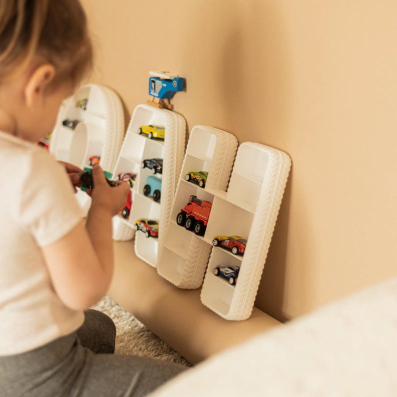 K&ouml;nnte beinhalten: Wei&szlig;e, buchstabenf&ouml;rmige Regale mit Spielzeugautos gef&uuml;llt. Die Regale bilden das Wort "DAH". Ein Kind spielt mit Spielzeugautos vor den Regalen. Die Regale sind an einer beigefarbenen Wand befestigt.