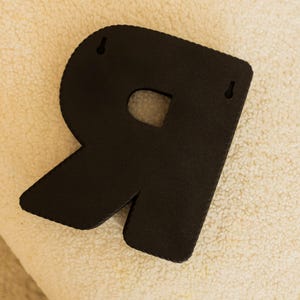 K&ouml;nnte beinhalten: Ein schwarzer, strukturierter Wandbehang mit dem Buchstaben "R". Der Buchstabe ist ausgeschnitten und hat eine abgerundete Oberseite und einen diagonalen Schenkel. Zwei kleine L&ouml;cher zum Aufh&auml;ngen sind sichtbar. Der Hintergrund ist eine cremefarbene, flauschige Oberfl&auml;che.